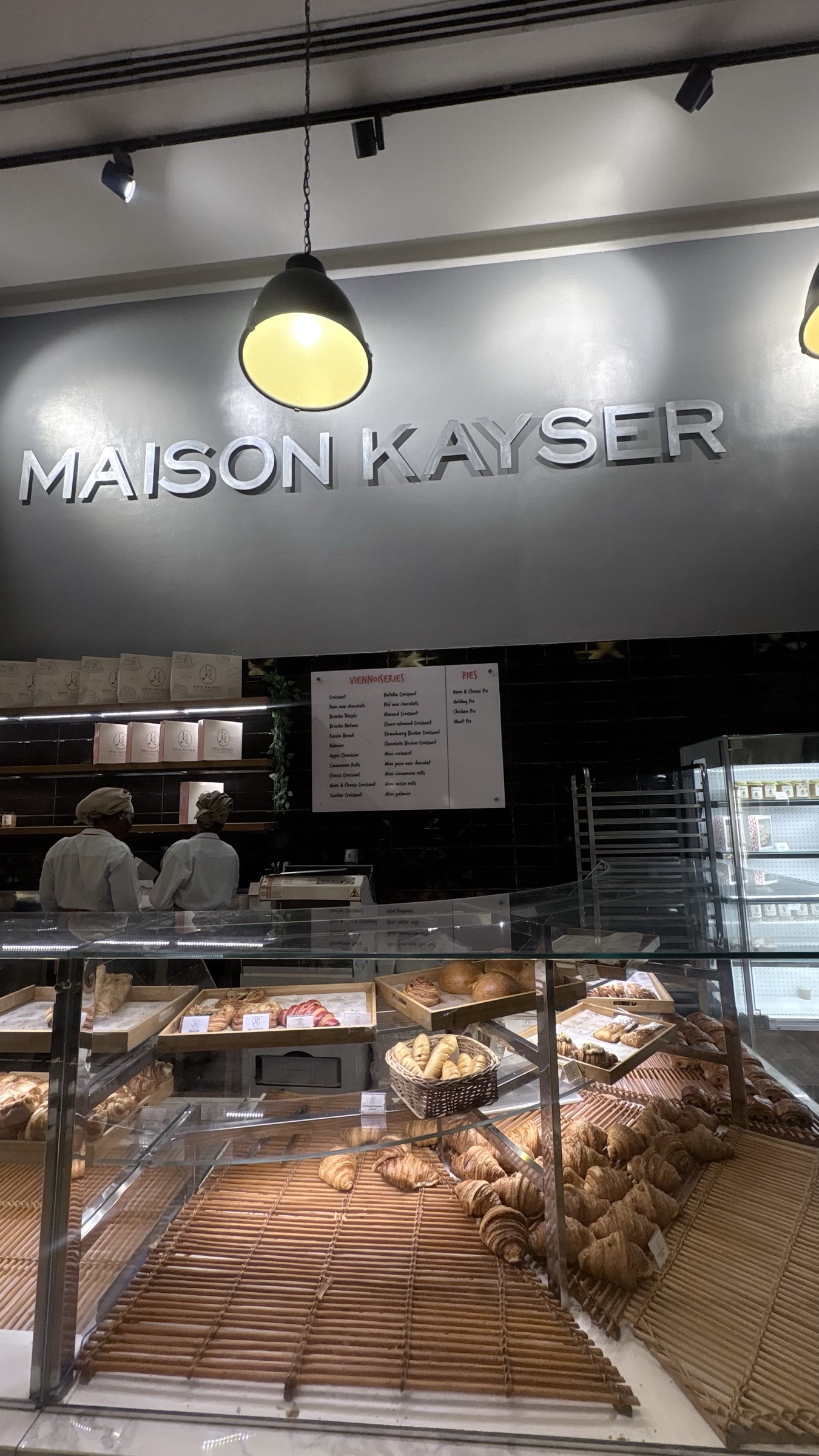 The Ikoyi bakery counter under the Maison Kayser sign, croissants and viennoiserie in the case and bakers at the pass