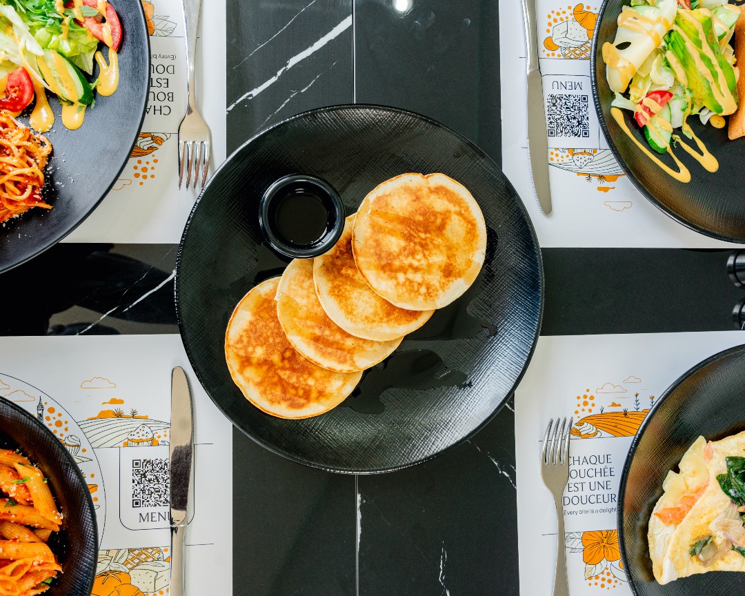 Pancakes at the centre of a shared table, surrounded by pasta, salads and illustrated Maison placemats